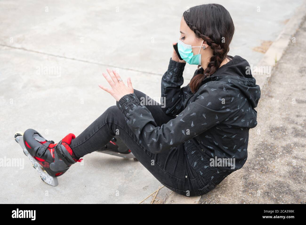 Woman in protective face mask on roller skating pause, sitting on the street and using mobile phone during coronavirus pandemic outbreak. Urban Girl Stock Photo