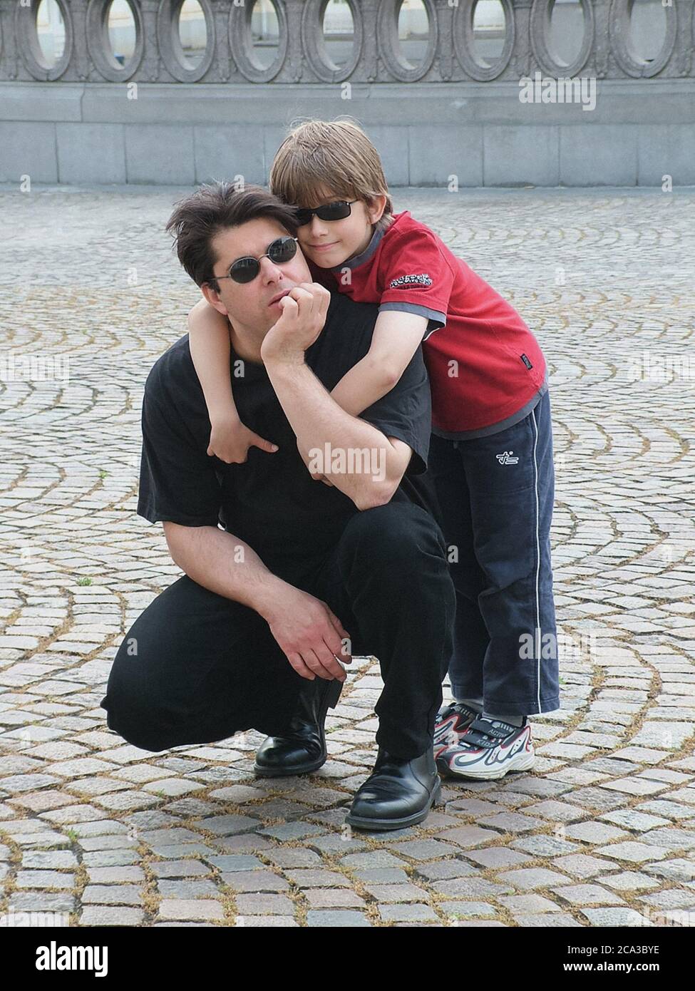 Boy and kneeling man both wearing black sunglasses hug on the fan shaped cobbles in front of an elipical stone balustrade in Belgium. Stock Photo