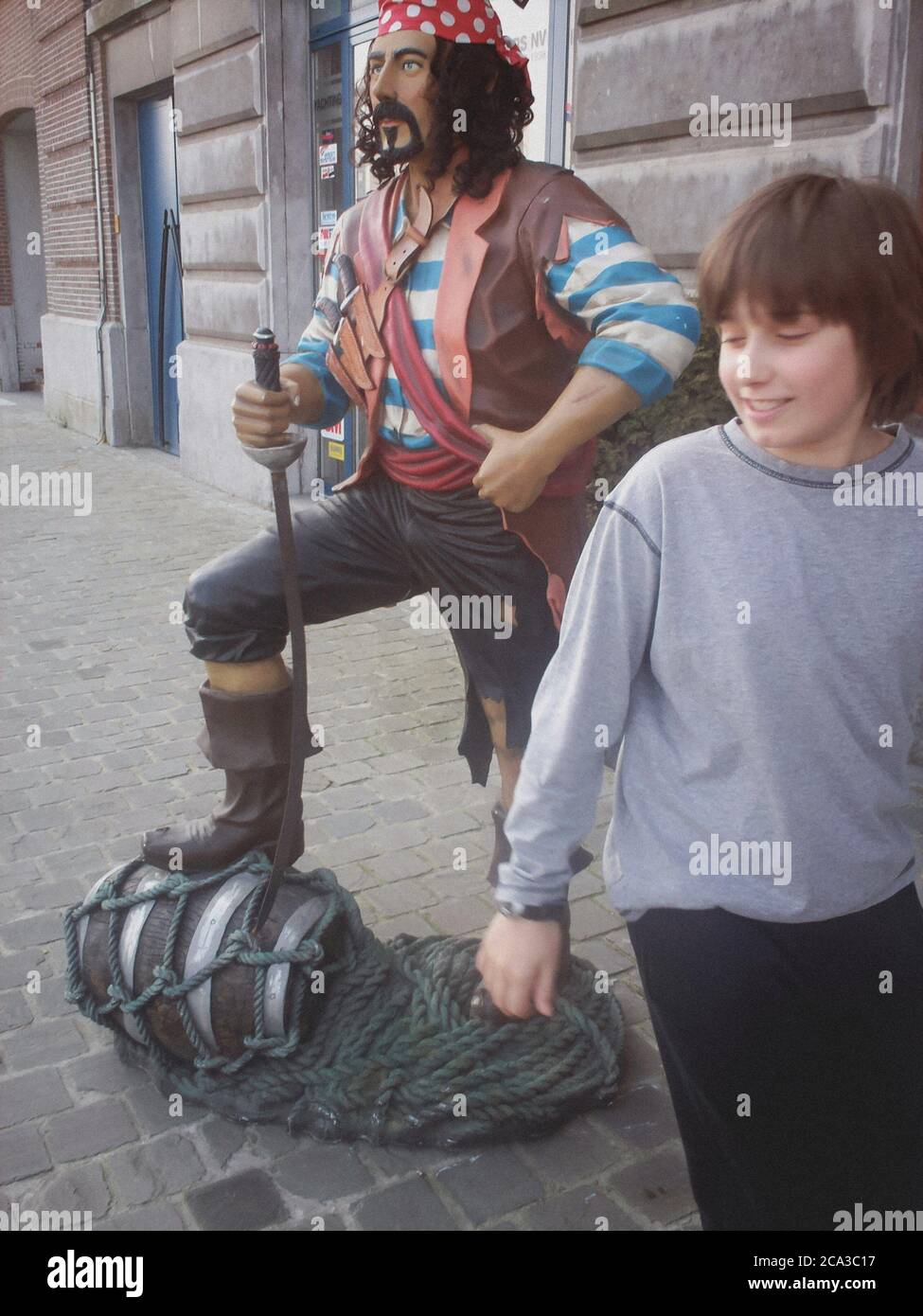Casually dressed young boy chuckles as he looks back at a flamboyant, brightly coloured model of a buccaneer in Antwerp, Belgium. Stock Photo