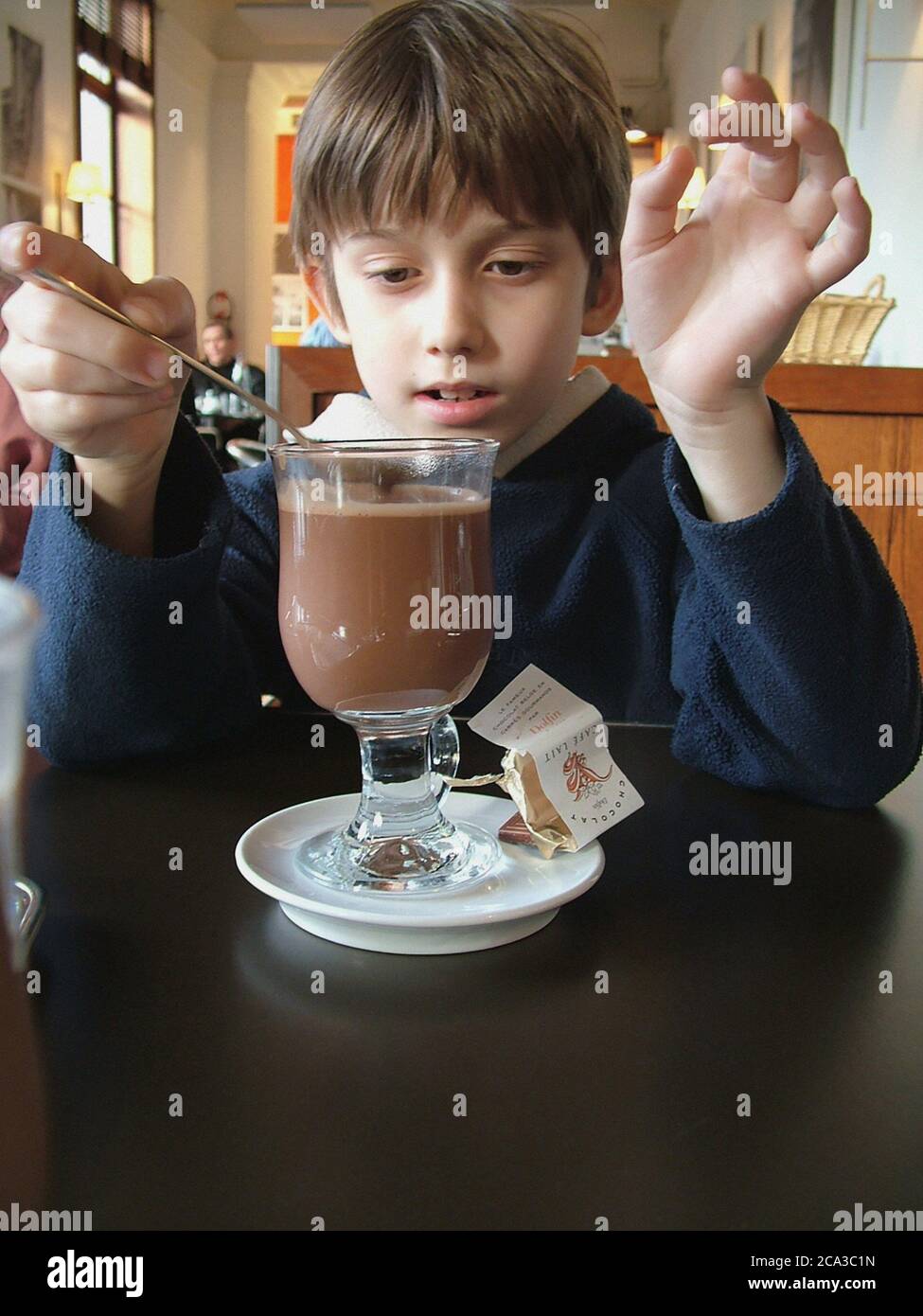 Young boy tastes frothy hot chocolate drink in a glass with chocolate sweet treat and signals perfection with his hands in the sunlit cafe of the Stock Photo