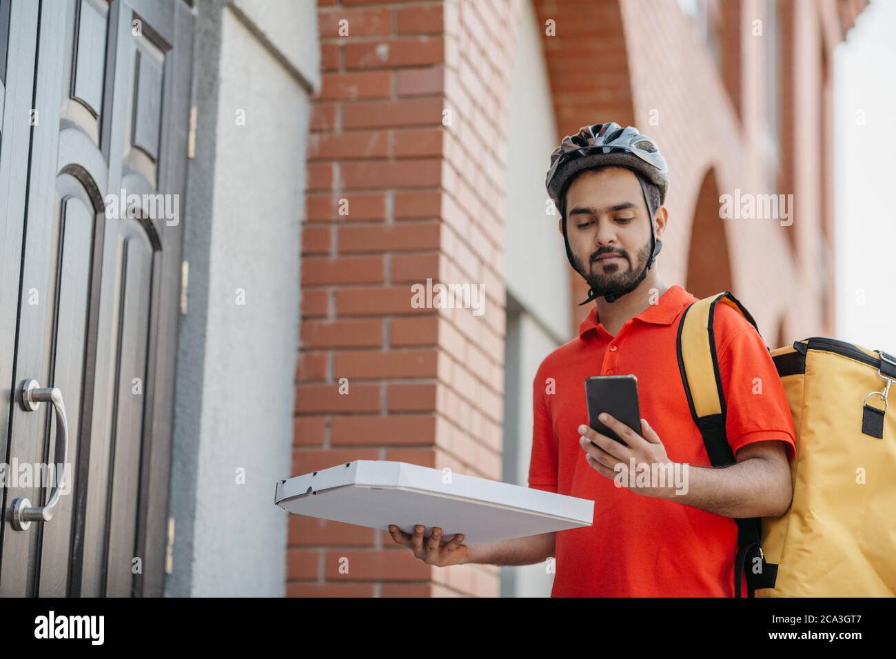 Fast delivery of pizza by courier in city. Serious man in helmet and bag, holds cardboard box with food and watches at smartphone Stock Photo