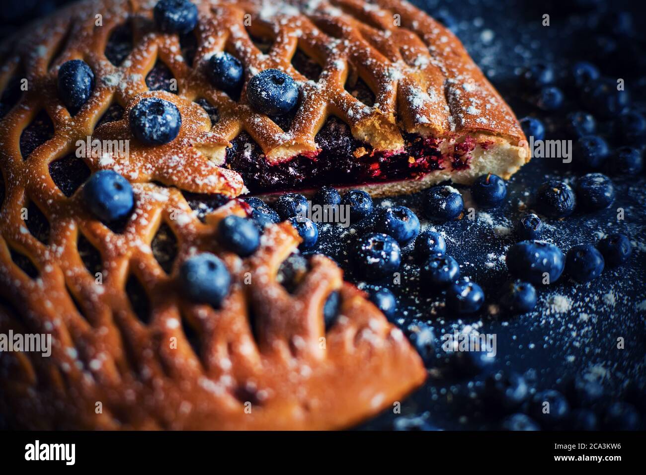 A round cut sweet pie with blueberry jam, next to which are ripe blue blueberries on a dark tray. Homemade cake. Stock Photo