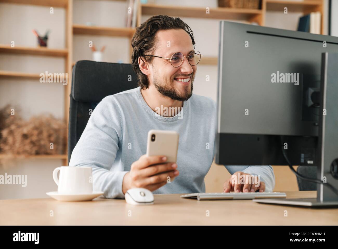 Image of smiling programmer man wearing eyeglasses using cellphone while working with computer in office Stock Photo