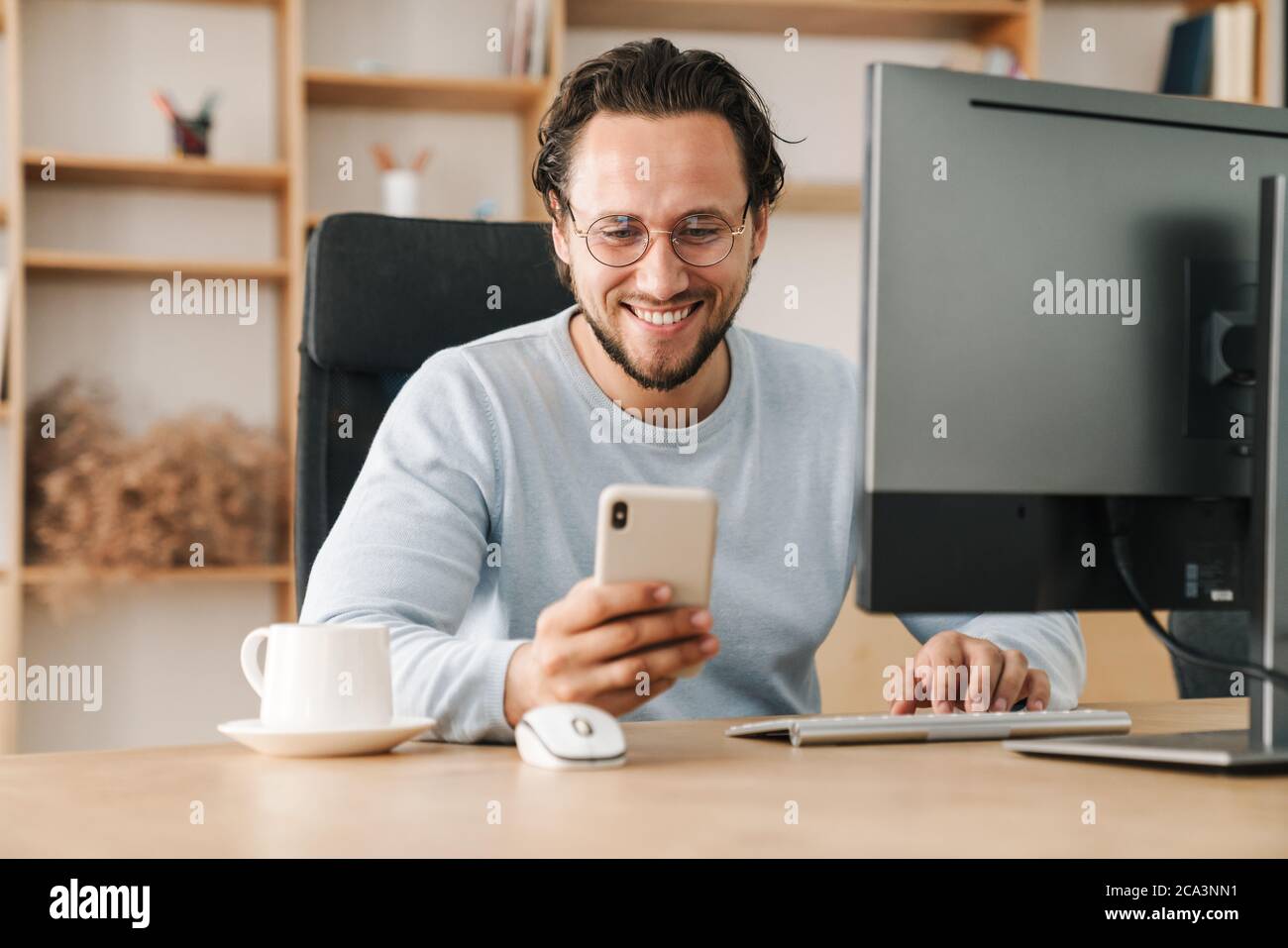 Image of smiling programmer man wearing eyeglasses using cellphone while working with computer in office Stock Photo
