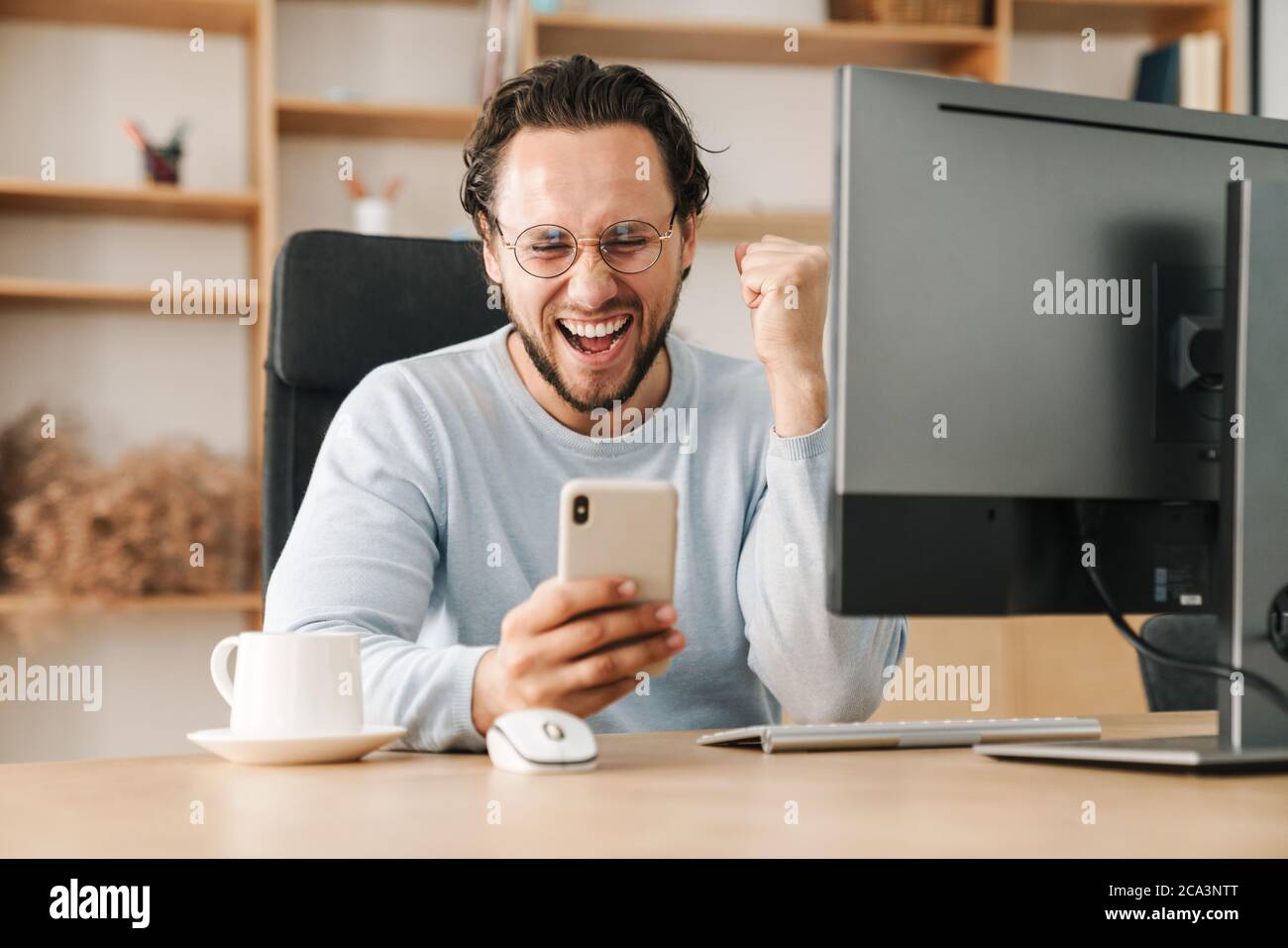 Image of excited programmer man making winner gesture and using cellphone while working in office Stock Photo