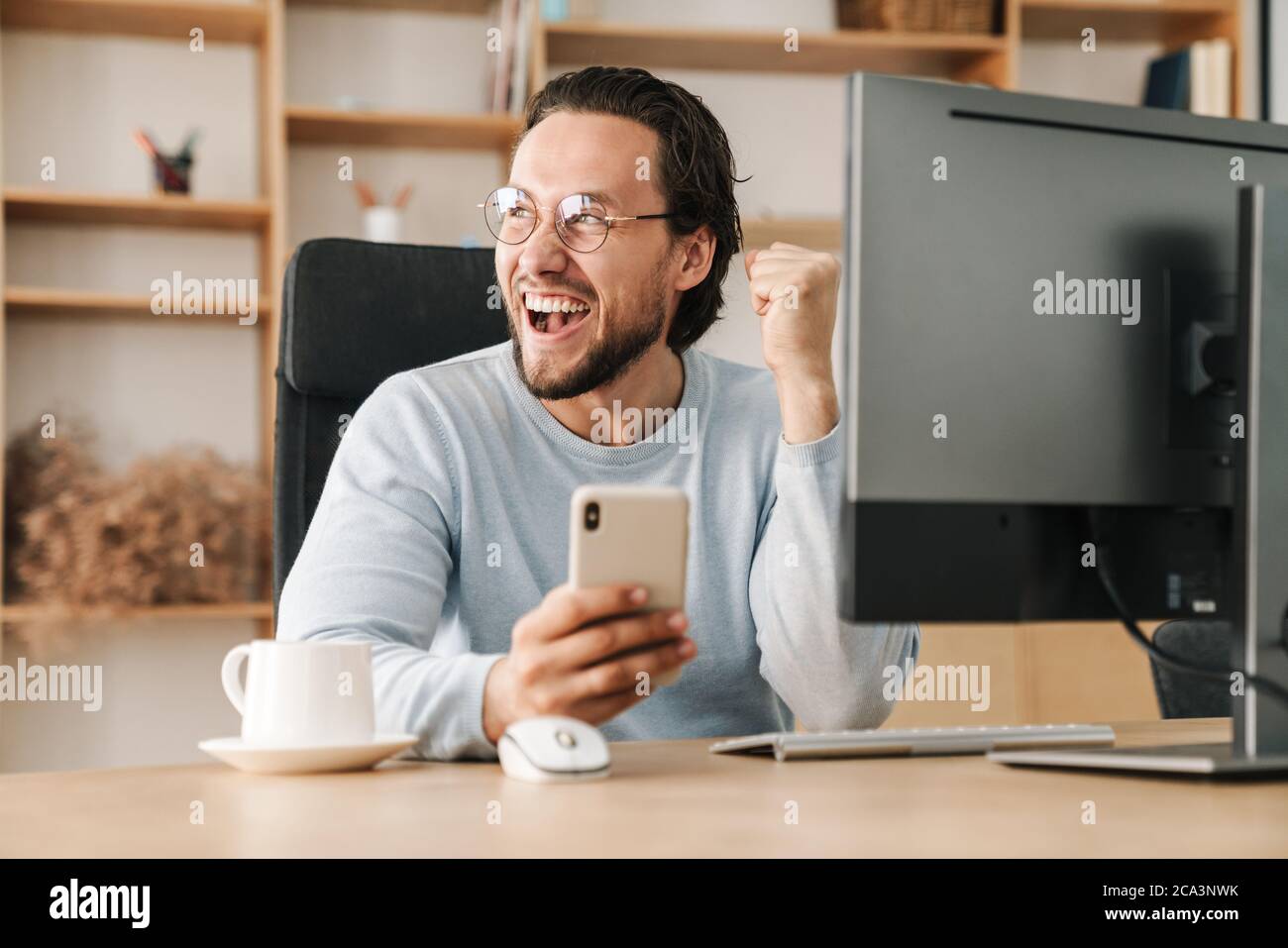 Image of excited programmer man making winner gesture and using cellphone while working in office Stock Photo
