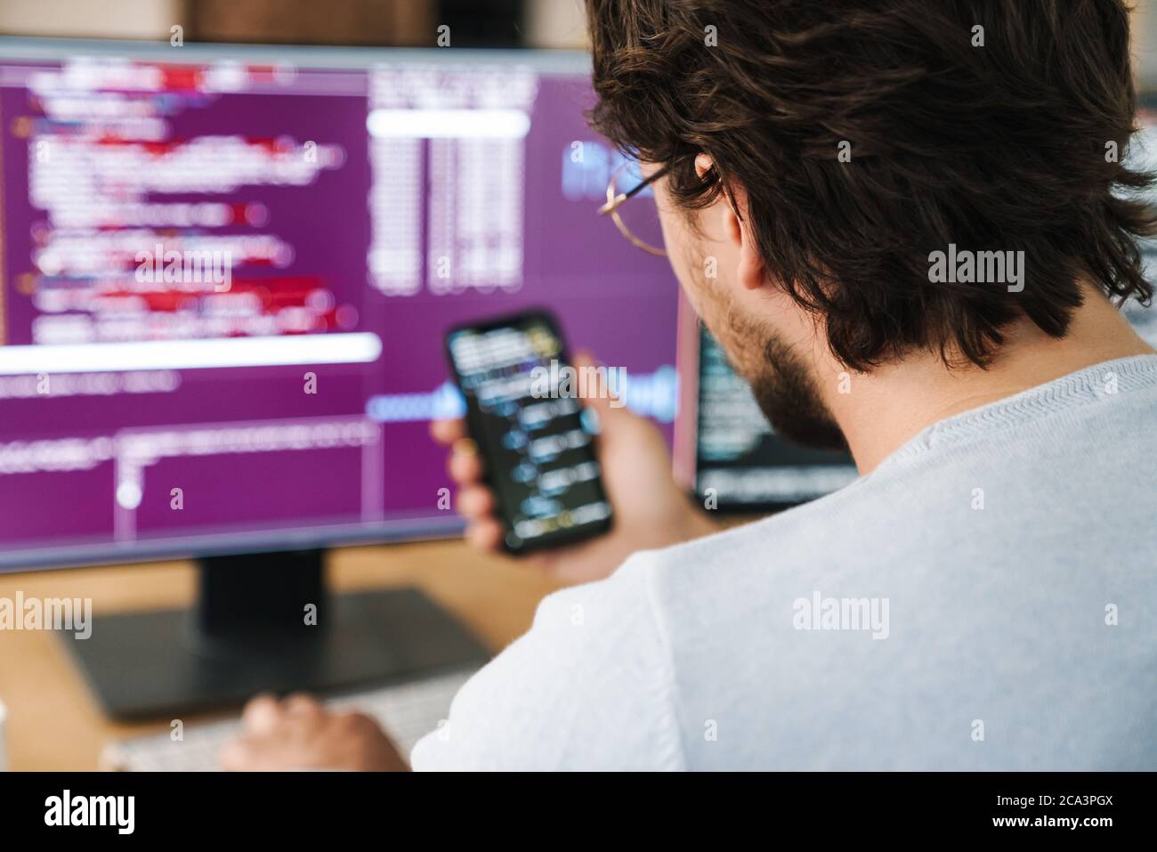 Image of unshaven focused programmer man using smartphone while working with computers in office Stock Photo