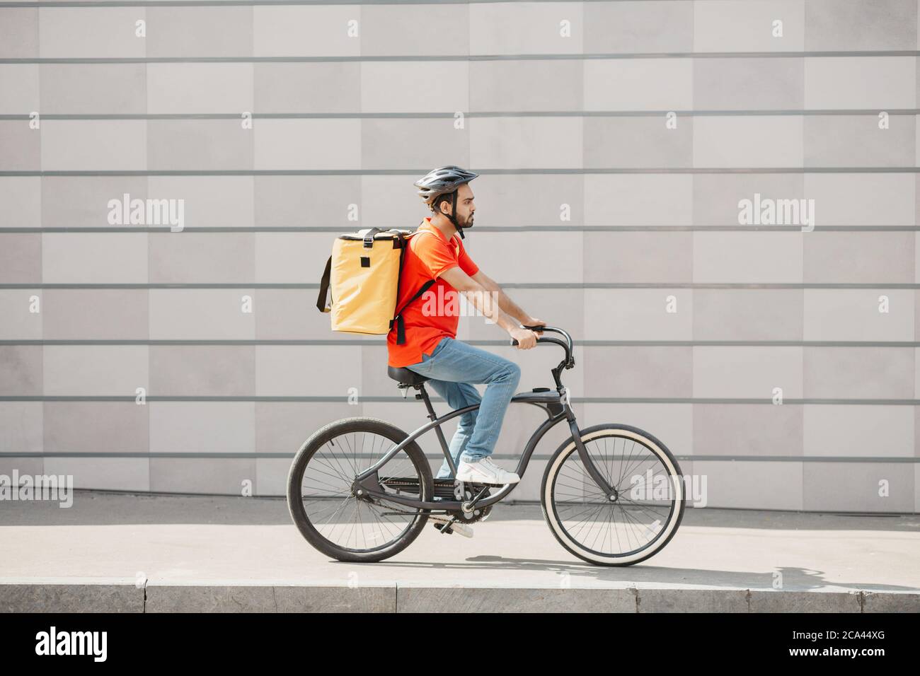 Courier cyclist on gray wall background. Young man in helmet with backpack rides on road Stock Photo