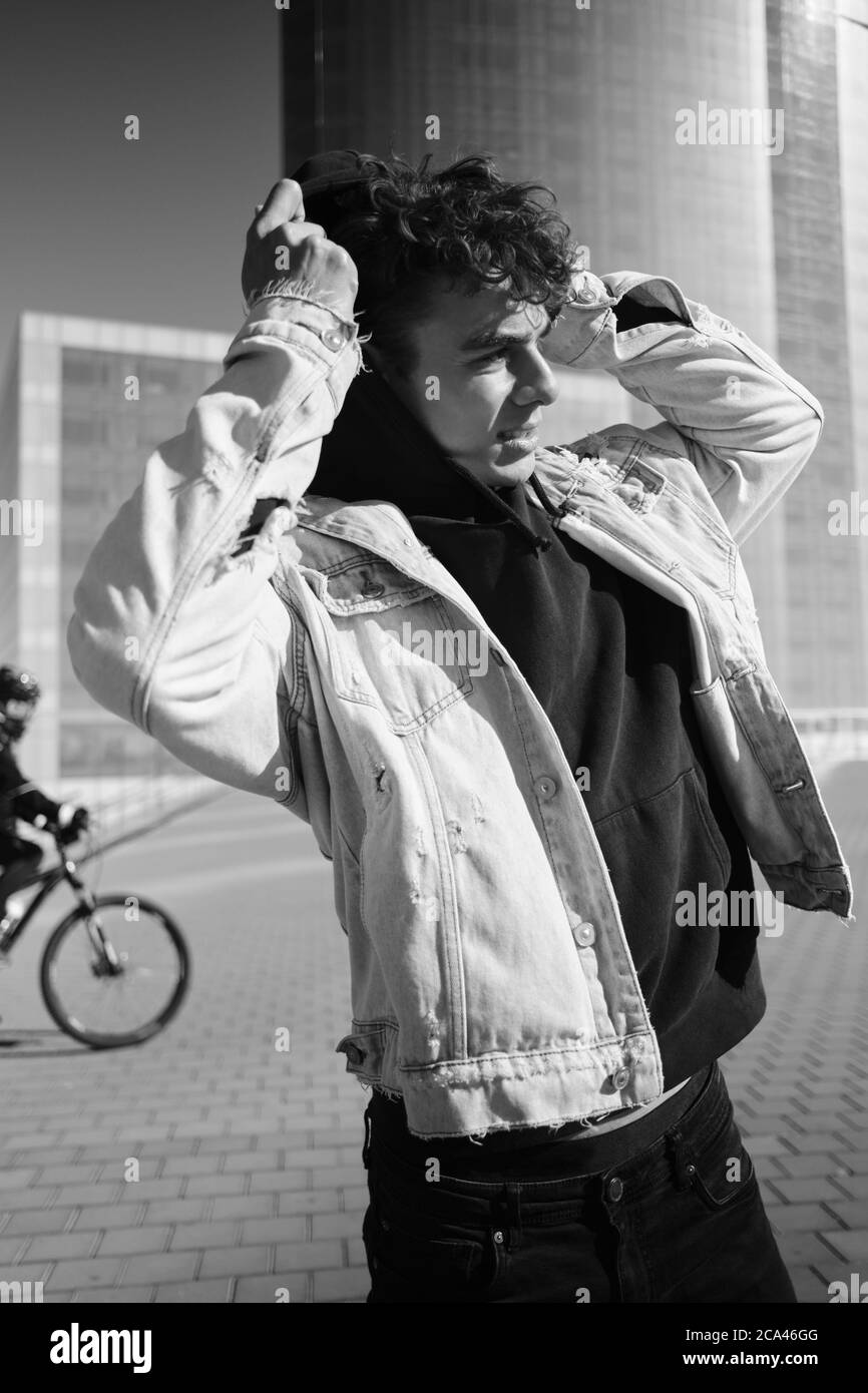 Handsome stylish guy wearing denim jacket with hoodie intently looking away on city street. Black and white photo Stock Photo
