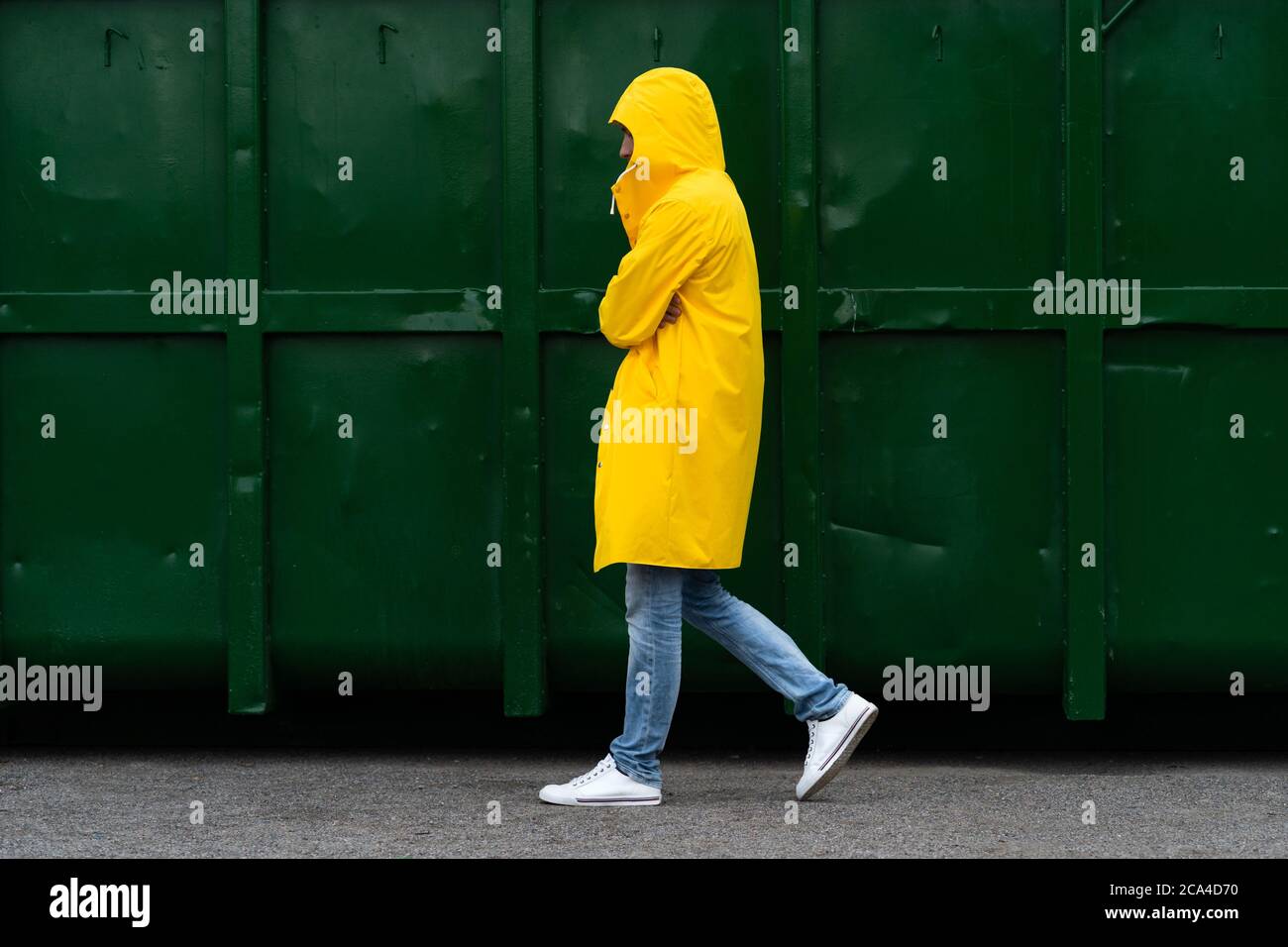 Man in a yellow raincoat walks down the street in the rain weather next to green container, side view. Outdoor. Stock Photo