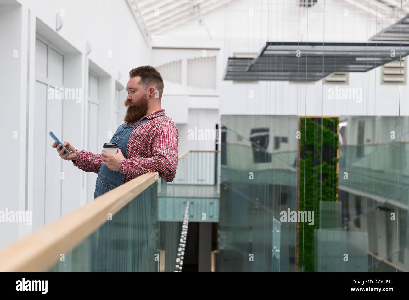 Brutal bearded self employed worker in blue overalls, checked shirt, drinking coffee from a paper cup, using smartphone. Time to take break. Hipster b Stock Photo