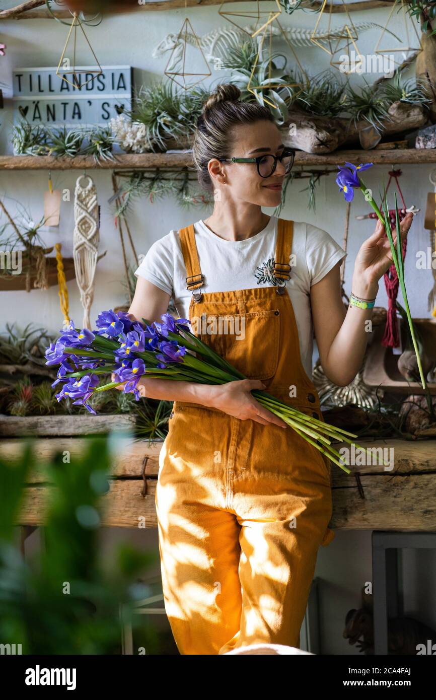 Surprised florist woman in glasses, wear orange overalls surrounded by collection of tillandsia air plants, charmed with pleasant gift, smiling, looki Stock Photo