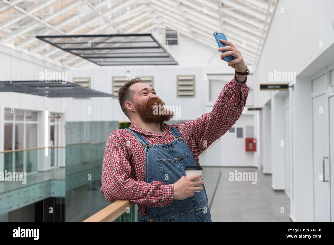 Smiling bearded barber worker in blue overalls, checked shirt, drinking coffee from a paper cup, talking on a video call or taking a selfie on smartph Stock Photo