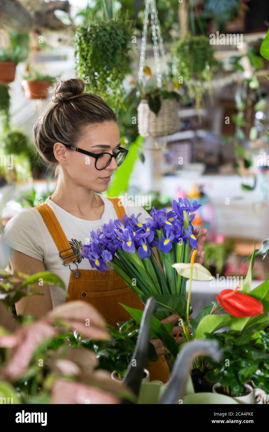 Surprised florist woman in glasses, wear orange overalls surrounded by houseplants, charmed with pleasant gift, looking at a bouquet of irises flowers Stock Photo