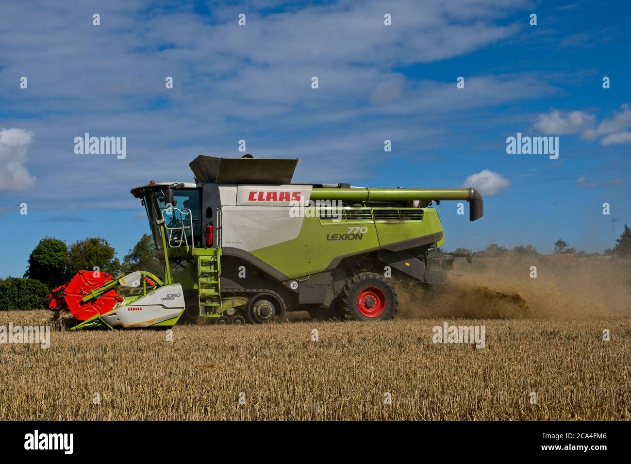 Winter Harvest Close side view of combined harvester at work in field Dusty Sunny cloudy sky Fields Trees and houses in distance Landscape format Stock Photo