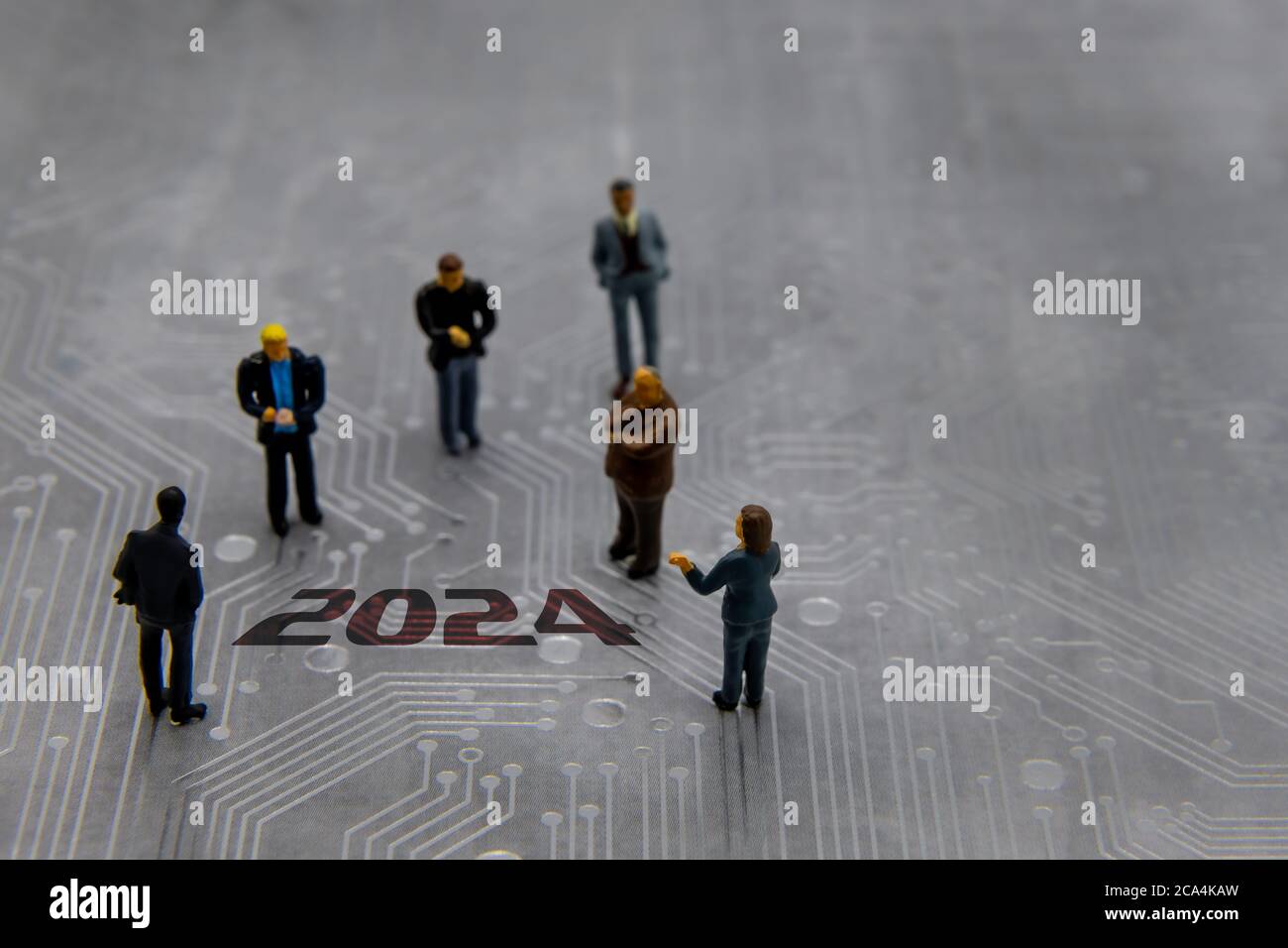 Miniature figurines posed as business people in a meeting over abstract futuristic circuit board with text year 2024 Stock Photo