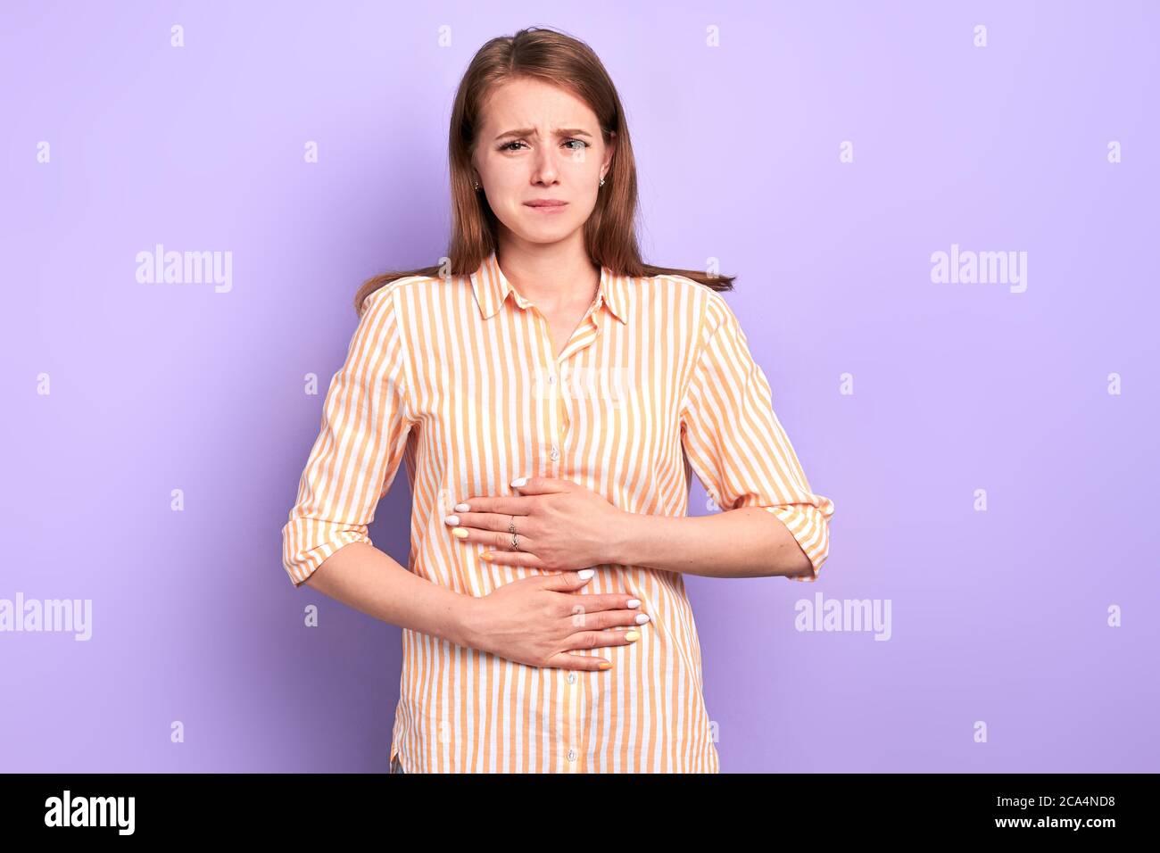 Sad young woman suffers from stomachache, feels discomfort after eating spoiled food, has painful face expression, hands on belly, dressed in casual s Stock Photo