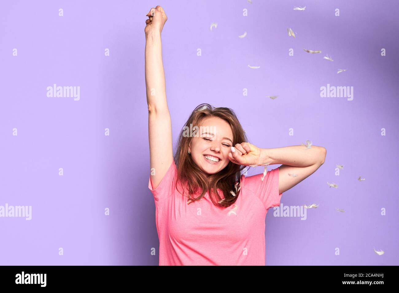 Front view of happy cute girl dancing under flying feathers with closed eyes during pillow fight party with her friends, feeling playful and joyful em Stock Photo