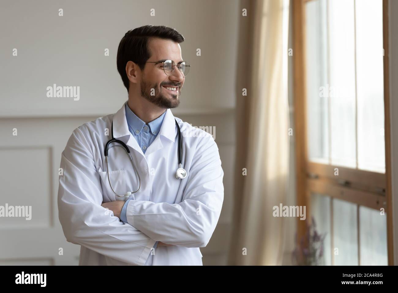 Smiling young doctor planning future career, standing near window. Stock Photo