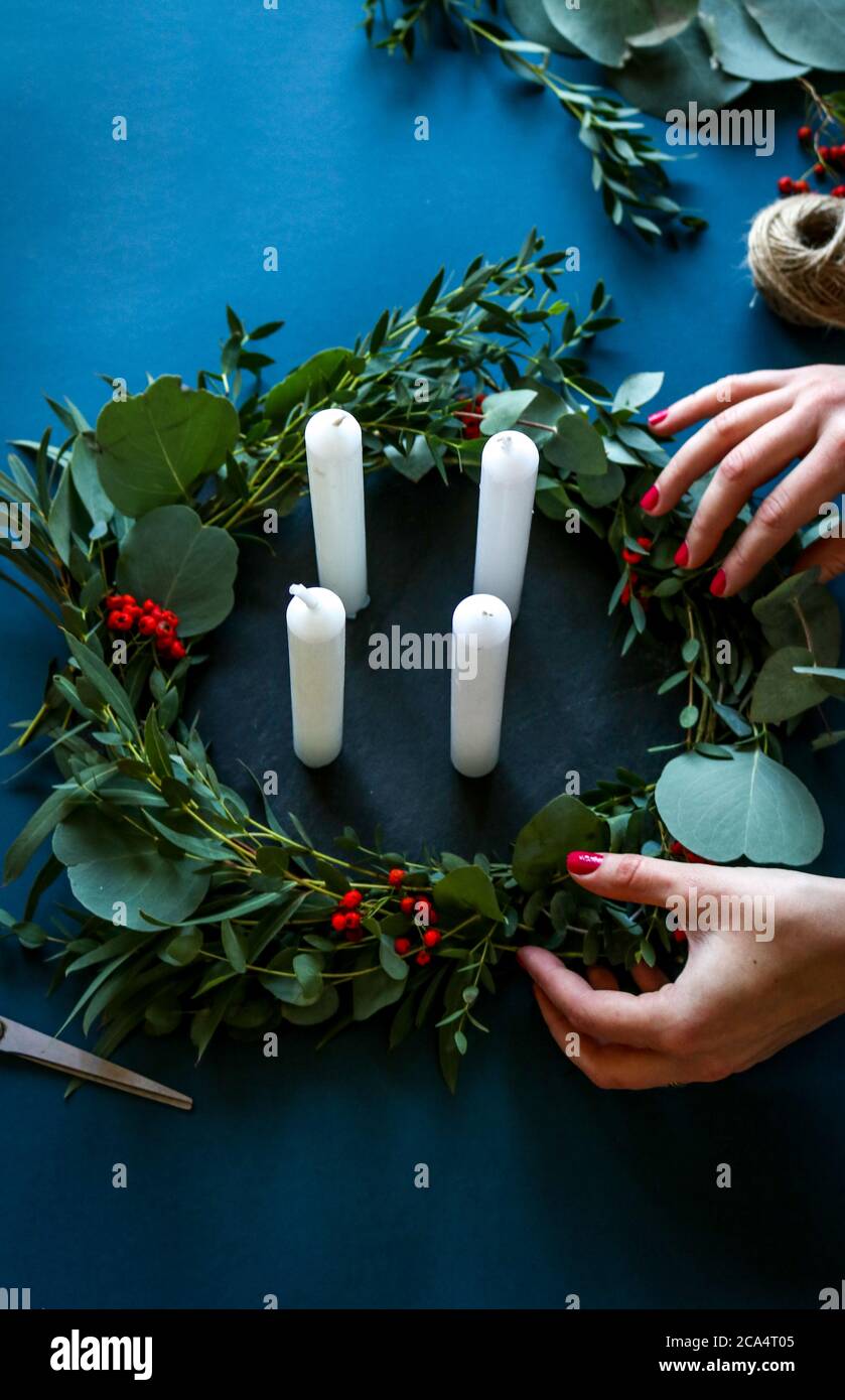 Woman making a modern Advent Christmas wreath with white candles, eucalyptus leaves and red berries on a blue background. Stock Photo