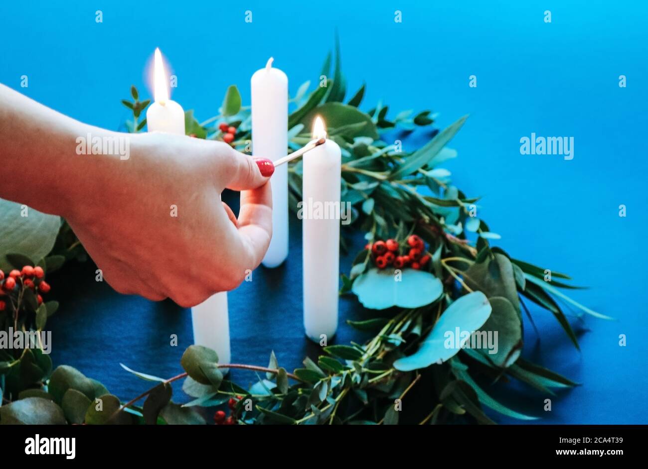 Close up of a woman lighting Advent candles on modern Christmas wreath with eucalyptus leaves isolated on blue coloured background Stock Photo