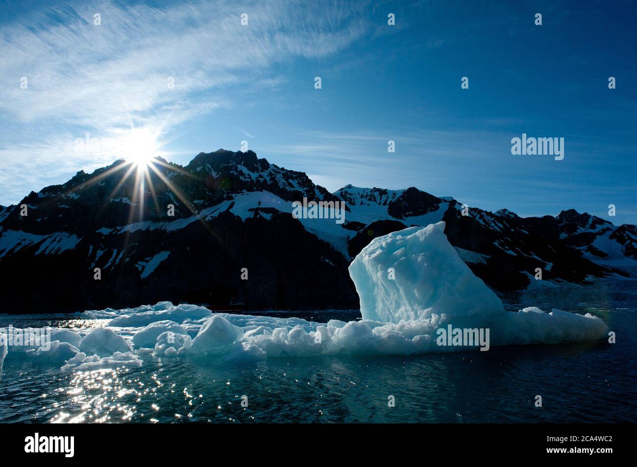 Backlit iceberg with sun starburst over snowy mountain as background shows lack of ice floes in fjord due to global warming climate crisis. Stock Photo