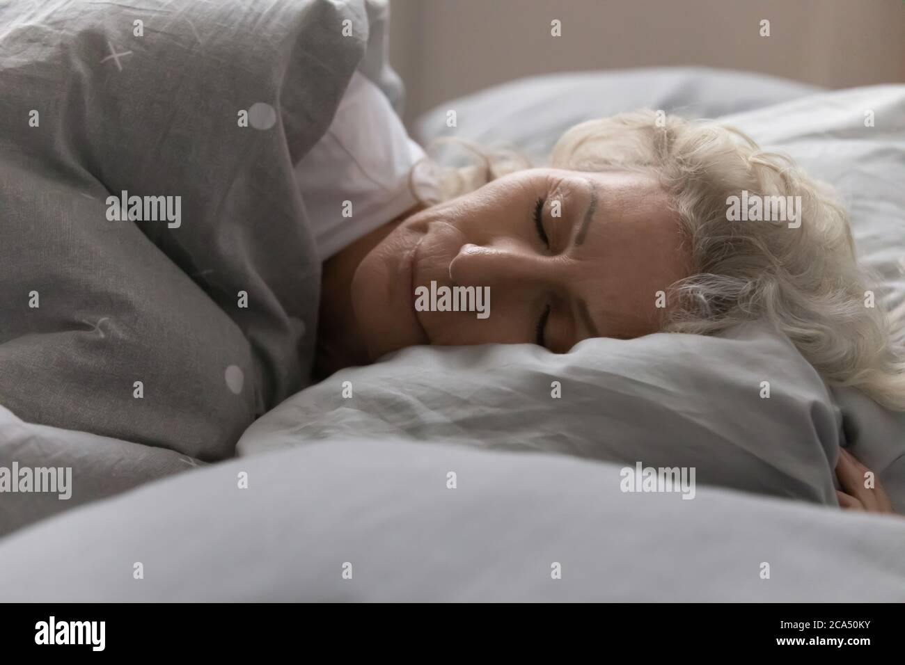 Elderly woman sleeping in bed under blanket in cozy bedroom Stock Photo