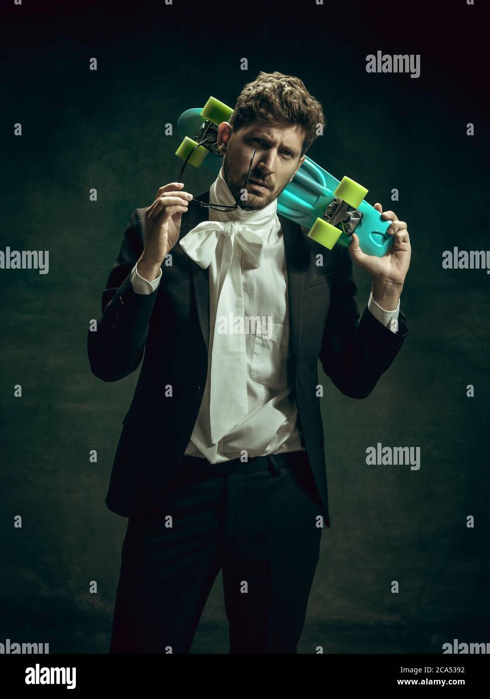 Skateboard urban style. Young man in suit as Dorian Gray isolated on dark green background. Retro style, comparison of eras concept. Beautiful male model like classic literature character, old-fashioned. Stock Photo