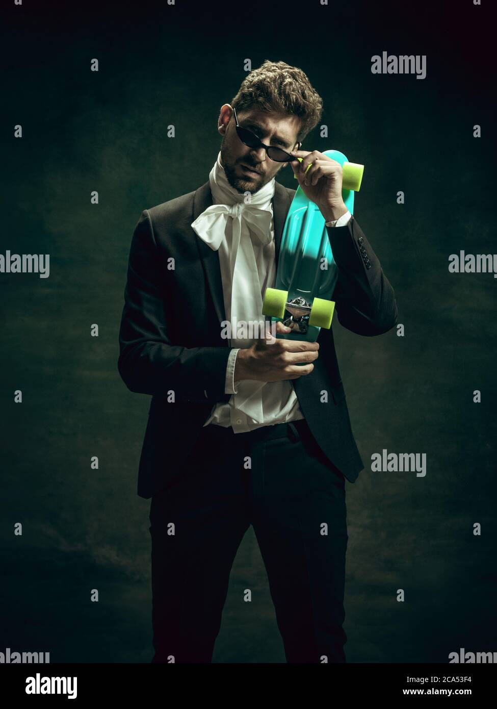 Skateboard urban style. Young man in suit as Dorian Gray isolated on dark green background. Retro style, comparison of eras concept. Beautiful male model like classic literature character, old-fashioned. Stock Photo