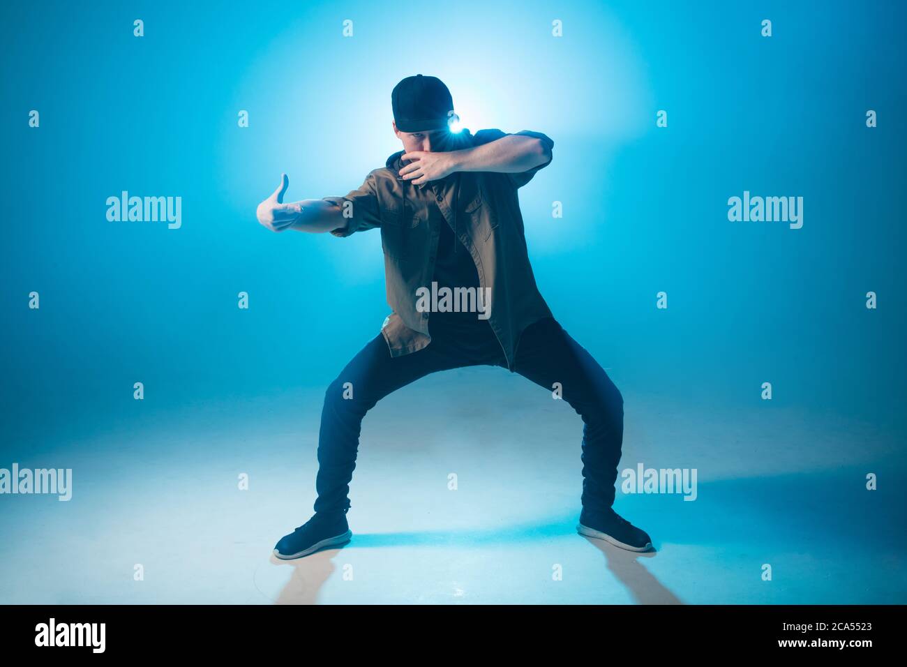Full length portrait of a young man dancer in casual street wear dancing funky hip hop on isolated studio neon blue background Stock Photo