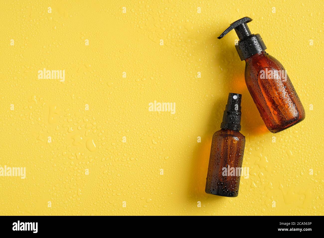 Amber glass cosmetic bottles in droplets of water. Refreshing beauty products on yellow background. Top view, flat lay. Stock Photo