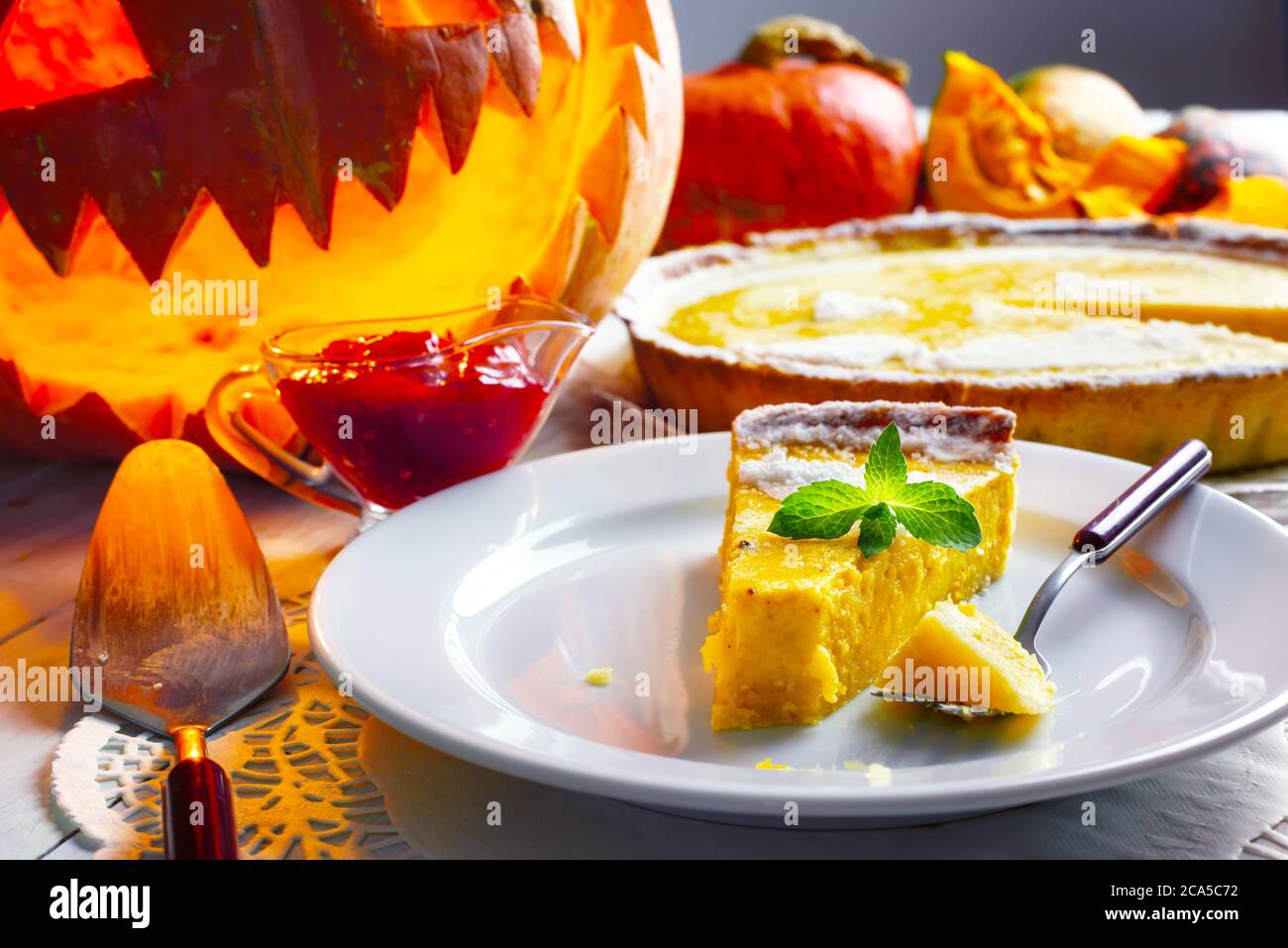 Piece of homemade halloween pumpkin cake with leaf of mint on white plate closeup. Jack-o-lantern, fork and cowberry jam around Stock Photo