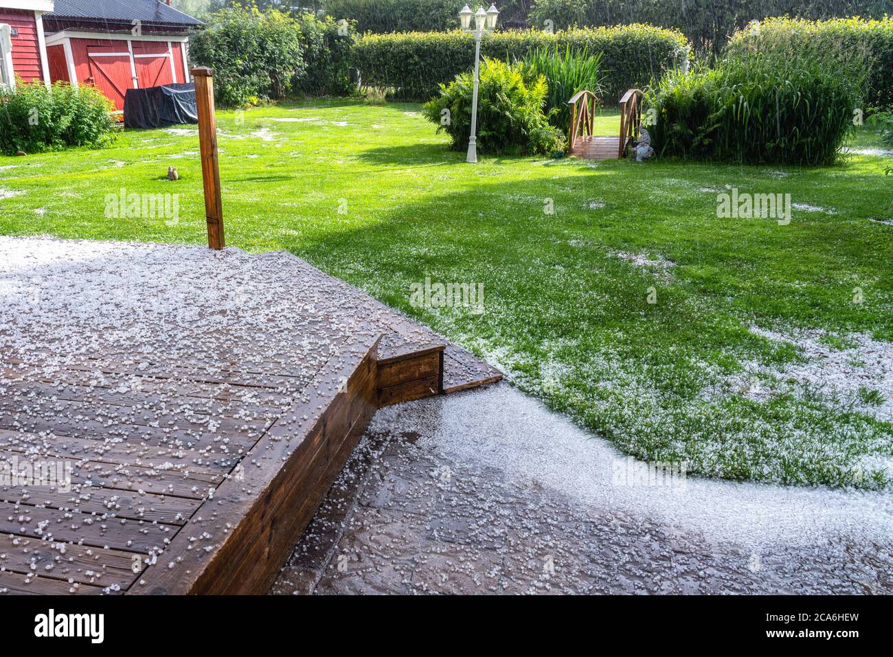 View at wooden terrace, stone path and green garden with hail stones during hailstorm from sky with sunlight Stock Photo
