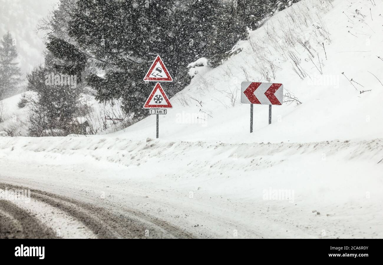 Sharp curve on road, with road signs (careful skid, snow) during heavy snowstorm blizzard in winter. Dangerous driving conditions. Stock Photo