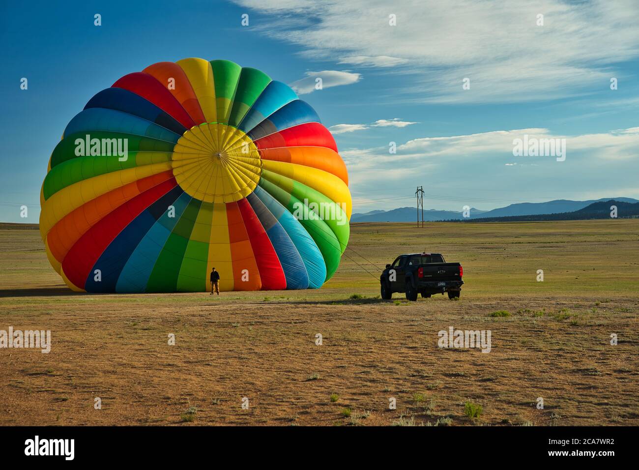 Hot air balloon beside man and truck shows the immense size of the large craft. Bright colors of the balloon contrast against the blue sky. Massive fl Stock Photo