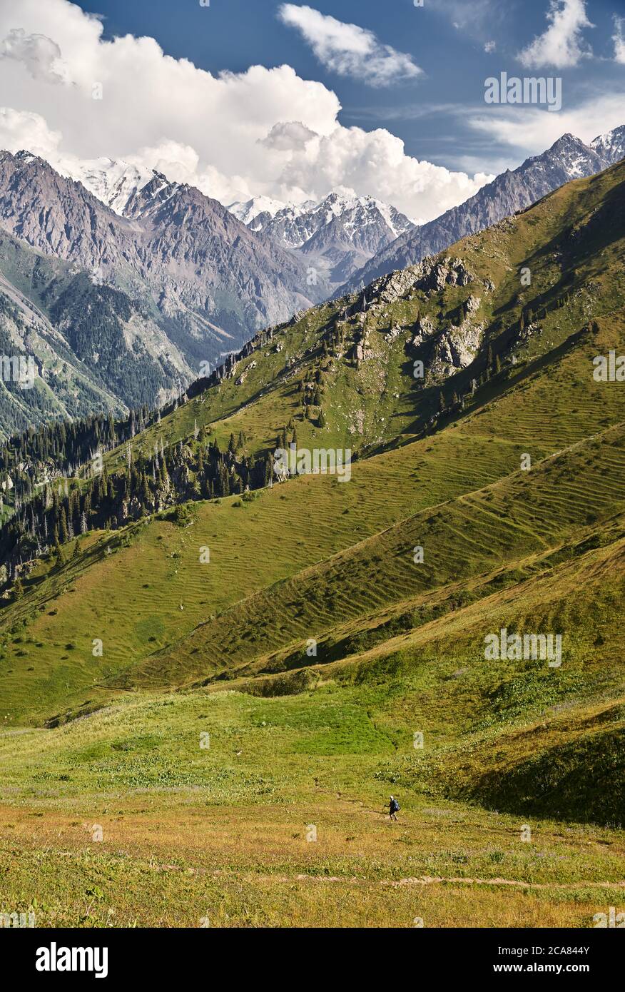 Small tourist walking on green hill in the mountain valley with high snowy peaks Stock Photo