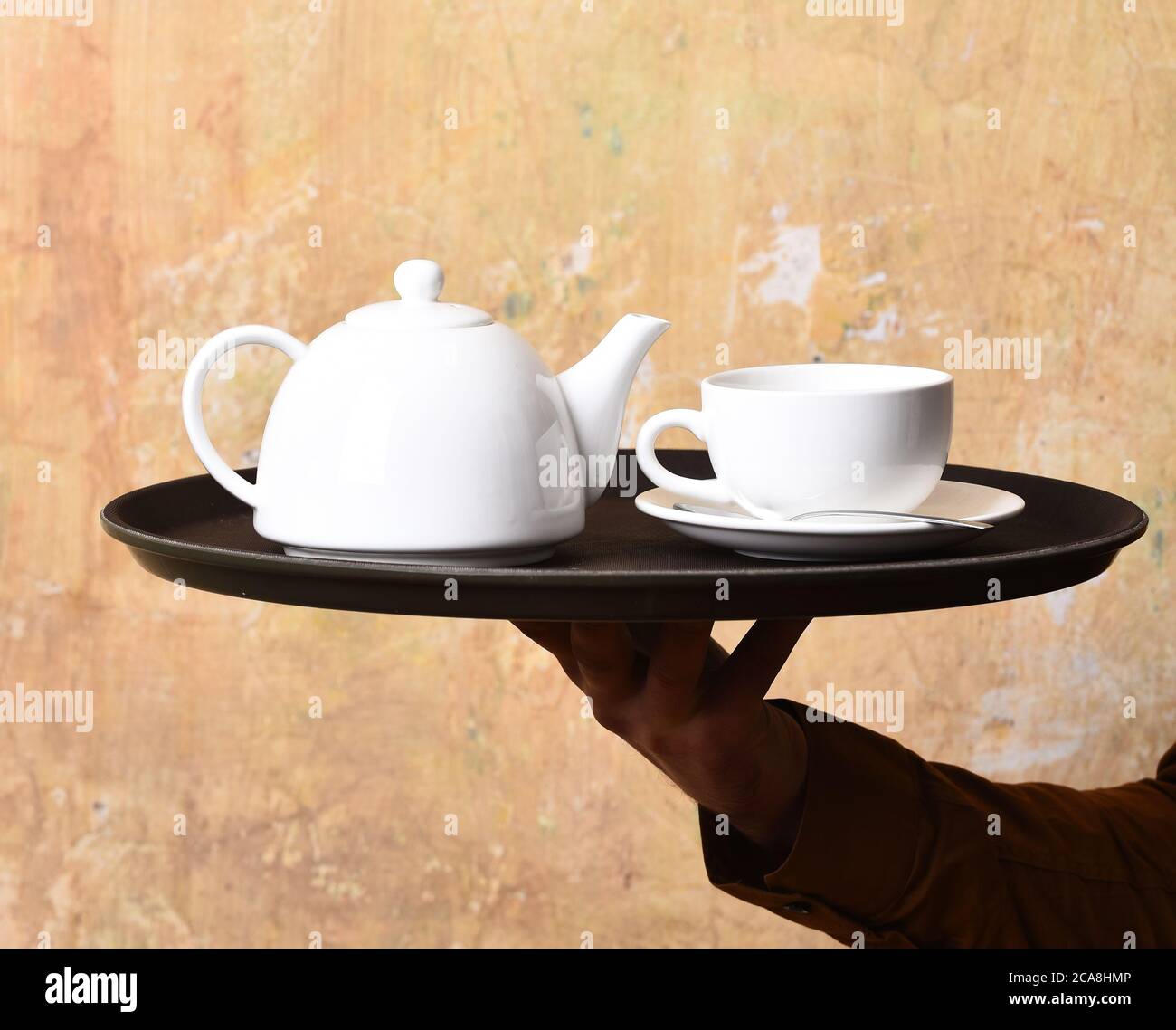 Barman brings tea or coffee. Service and restaurant catering concept. Waiters hand with white tea cup and pot on tray. Male hand serves tea on beige wall background, close up. Stock Photo