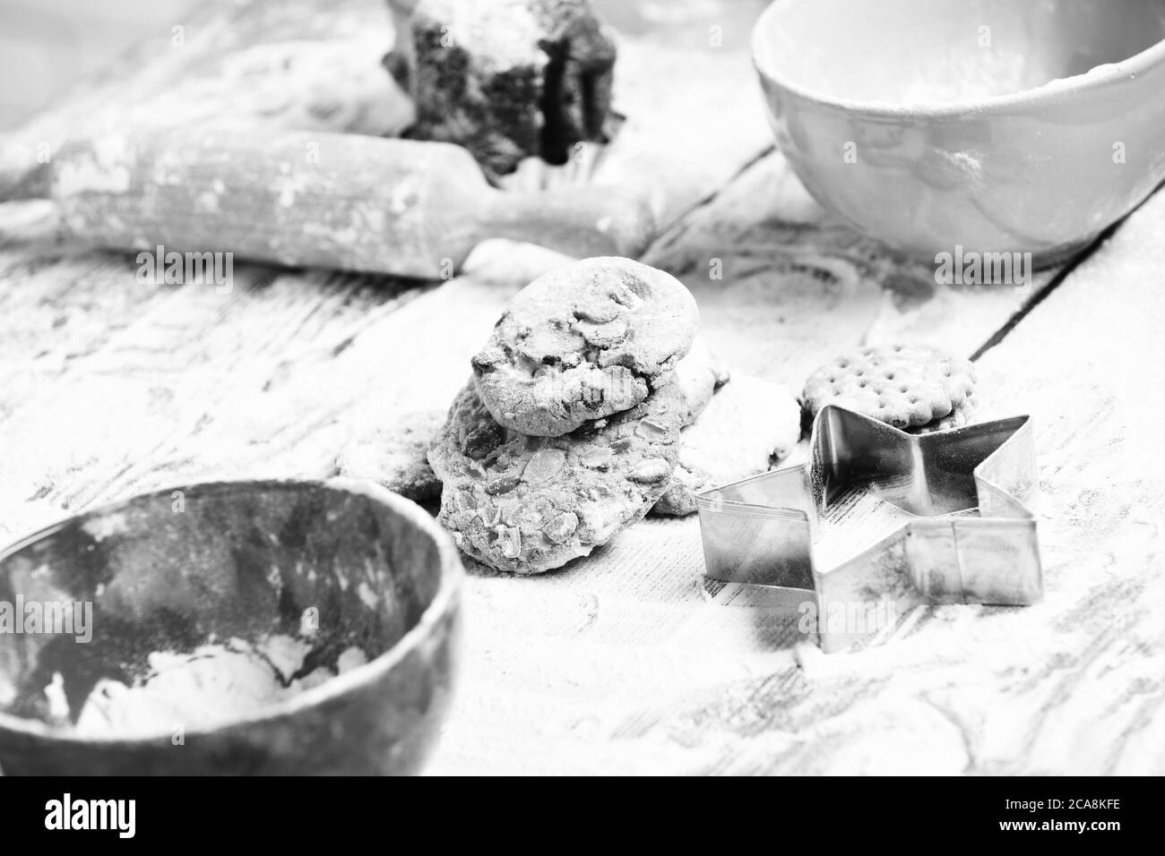tasty delicious chip chocolate cookies sprinkled with powdered sugar near red turquoise bowls, cutter and rolling pin on wooden table with lot of flour, selective focus Stock Photo