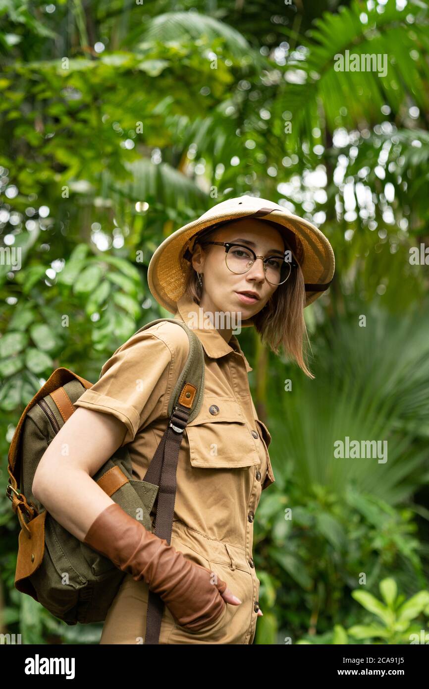 Woman botanist dressed in safari style in greenhouse. Naturalist in khaki clothes, leaver gloves with backpack walks in the rainforest surrounded by p Stock Photo