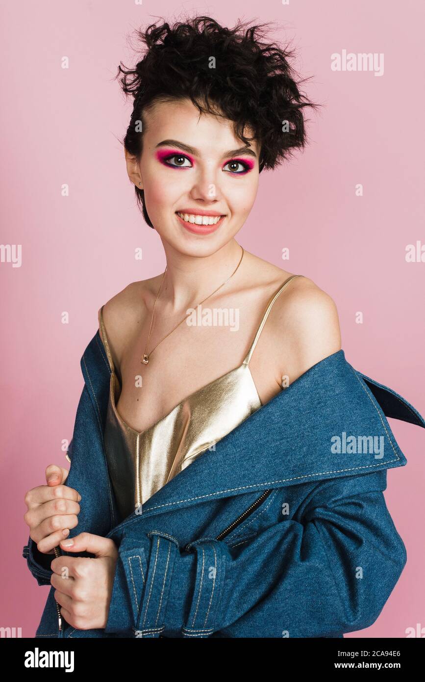 A girl with a sincere wide smile stands in the studio on a pastel pink background. A girl with a strange flying hairstyle and bright makeup Stock Photo