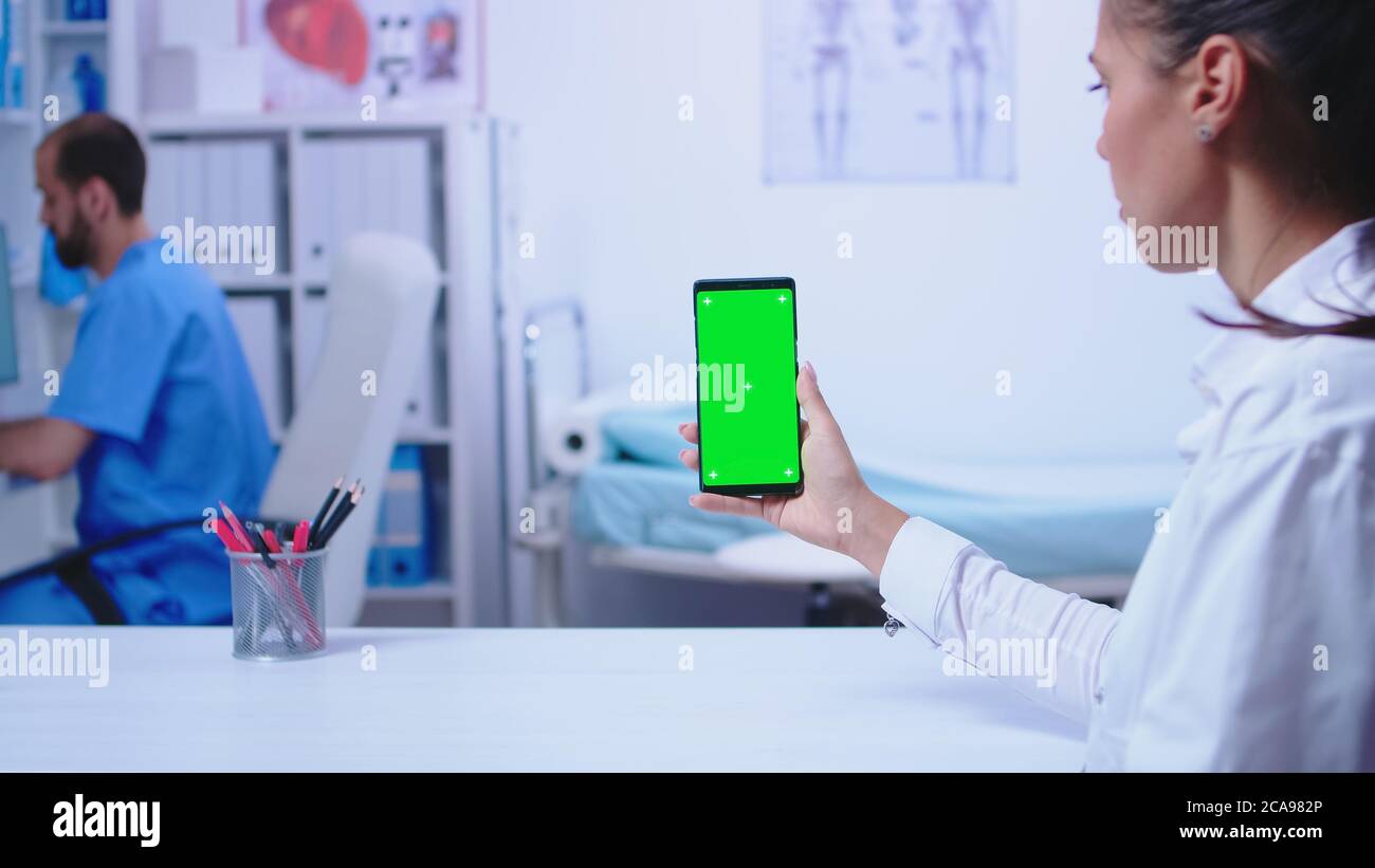 Medic in white coat holding smartphone with green screen in hospital