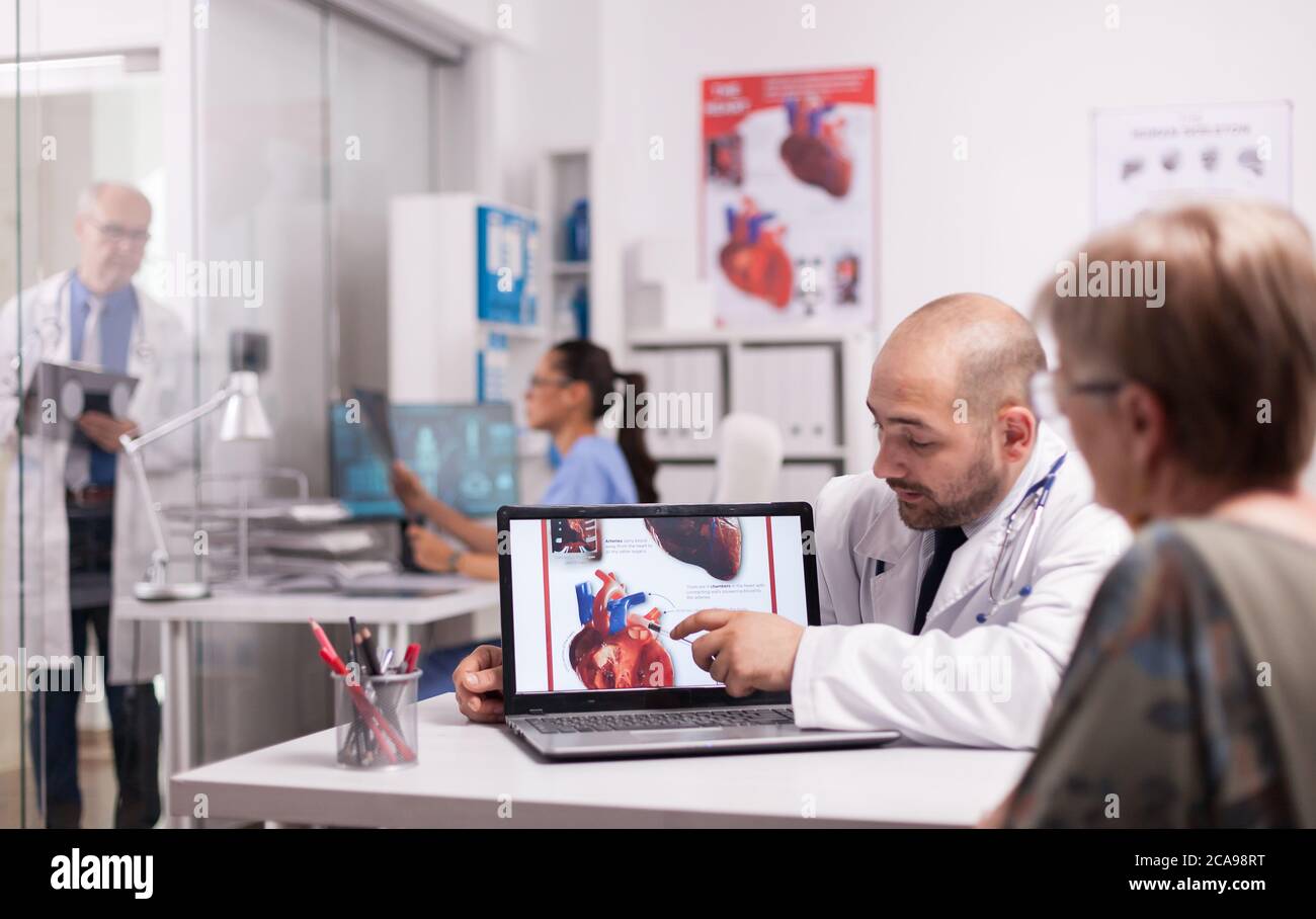 Young doctor pointing at heart on laptop screen in hospital office. Cardiologist with senior patient during consultation. Mature medic taking notes on clipboard on clinic corridor. Stock Photo