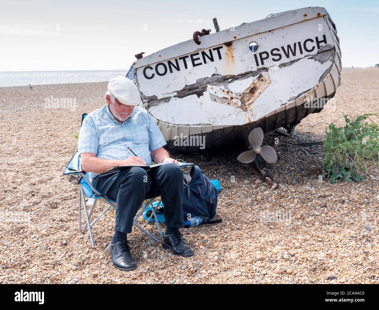 Aldeburgh, Suffolk, UK. 5th Aug, 2020. An artist is content to sit and draw on the shingle beach by the North Sea. It was a warm summer day with a stiff breeze as the current hot spell continues in the East of England. Credit: Julian Eales/Alamy Live News Stock Photo