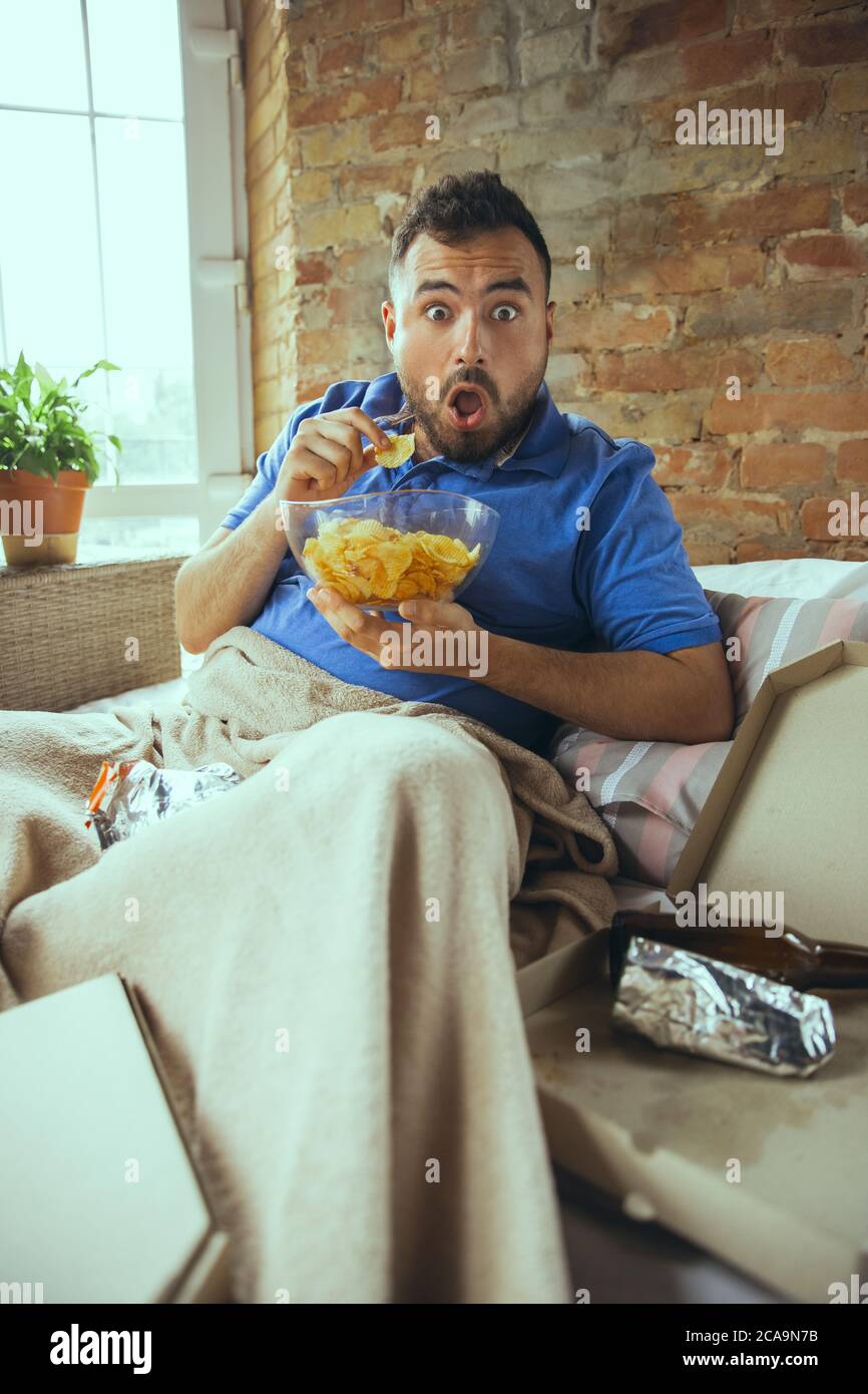 Shocked, biting crisps. Lazy man living in his bed surrounded with messy. No need to go out to be happy. Using gadgets, watching movie and series, looks emotional. Eating snacks and fast food. Stock Photo