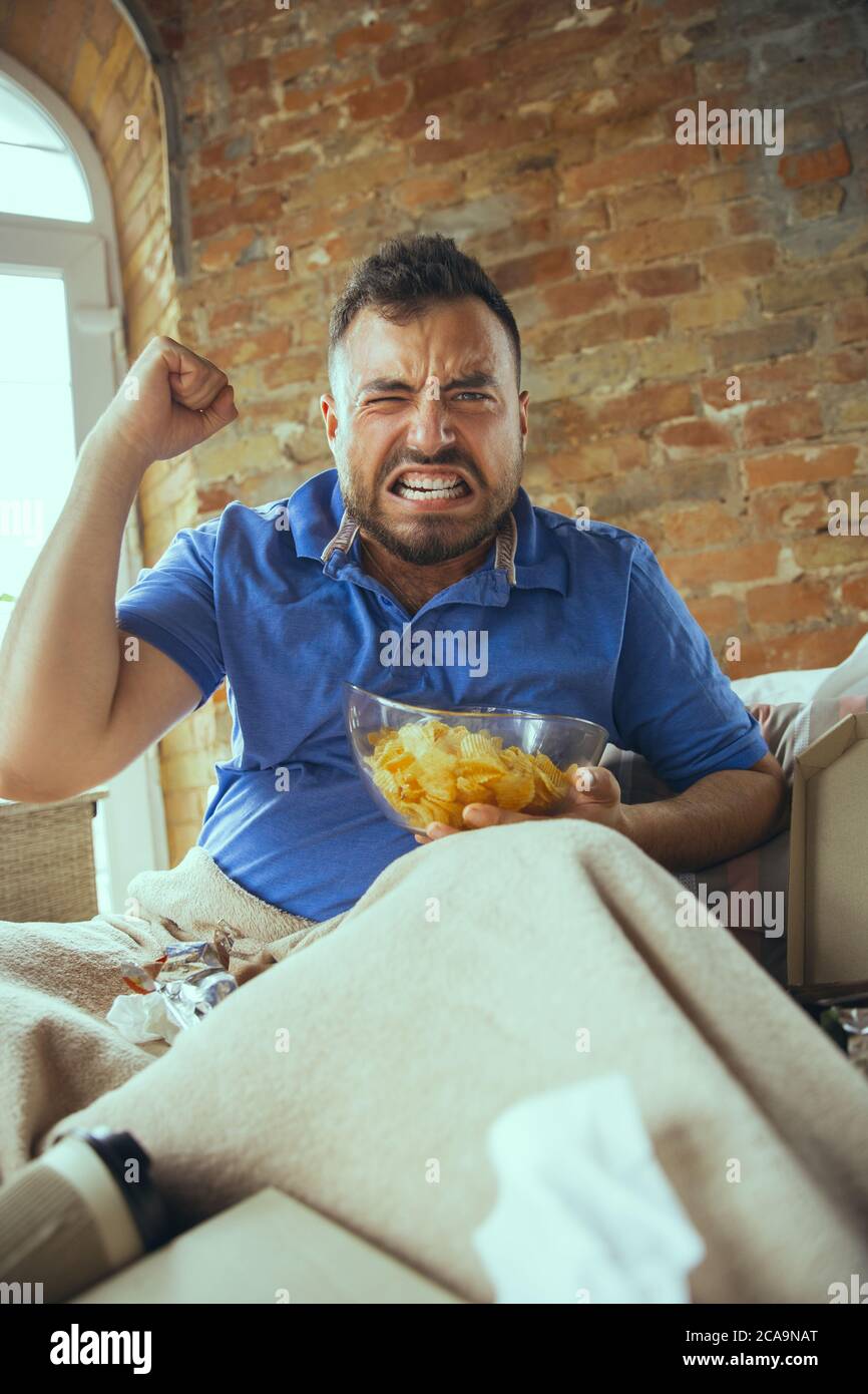 Sport fan cheering. Lazy man living the whole life in his bed surrounded with messy. No need to go out to be happy. Using gadgets, watching movie and series, looks emotional. Eating snacks and fast food. Stock Photo