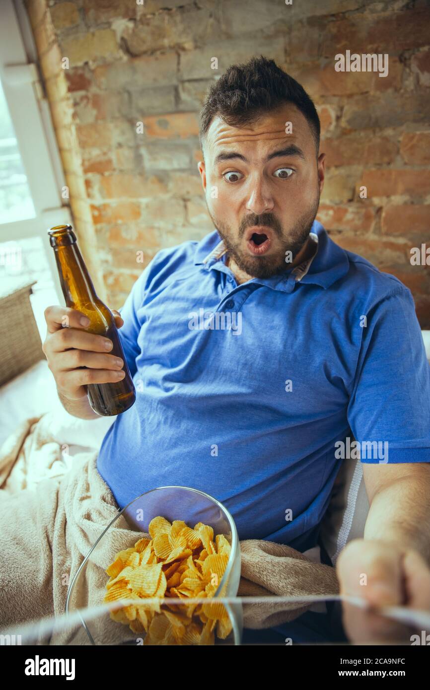Shocked, drinks beer. Lazy man living in his bed surrounded with messy. No need to go out to be happy. Using gadgets, watching movie and series, looks emotional. Eating snacks and fast food. Stock Photo