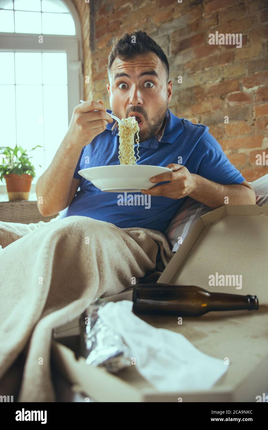 Shocked eating instant noodle. Lazy man living in his bed surrounded with messy. No need to go out to be happy. Using gadgets, watching movie and series, looks emotional. Eating snacks and fast food. Stock Photo