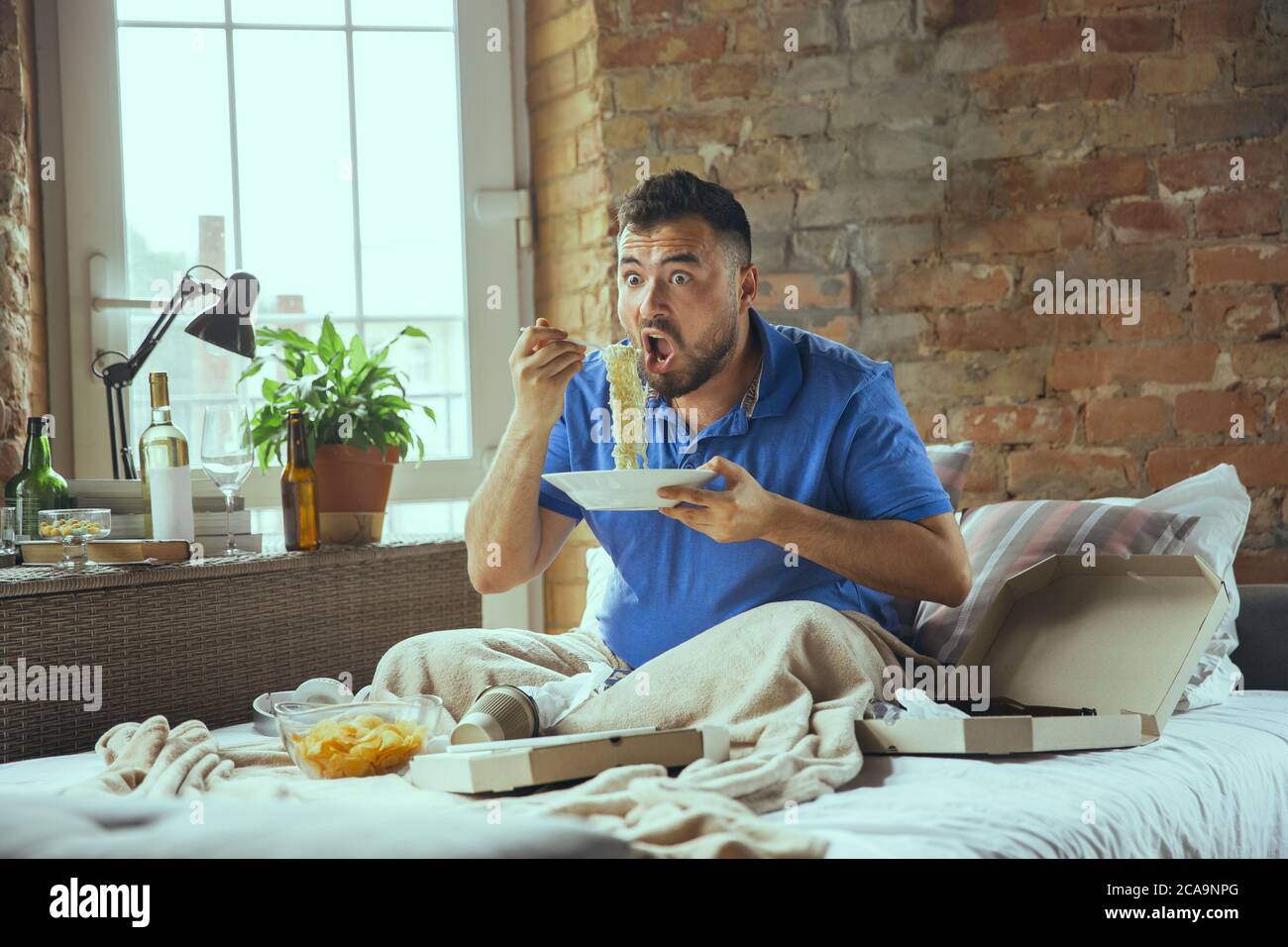 Shocked eating instant noodle. Lazy man living in his bed surrounded with messy. No need to go out to be happy. Using gadgets, watching movie and series, looks emotional. Eating snacks and fast food. Stock Photo