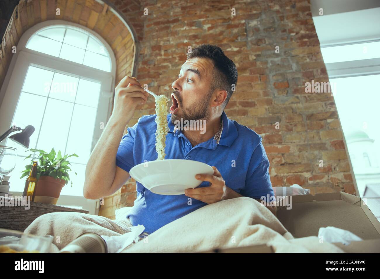 Passioned eating instant noodle. Lazy man living in his bed surrounded with messy. No need to go out to be happy. Using gadgets, watching movie and series, looks emotional. Eating snacks and fast food. Stock Photo