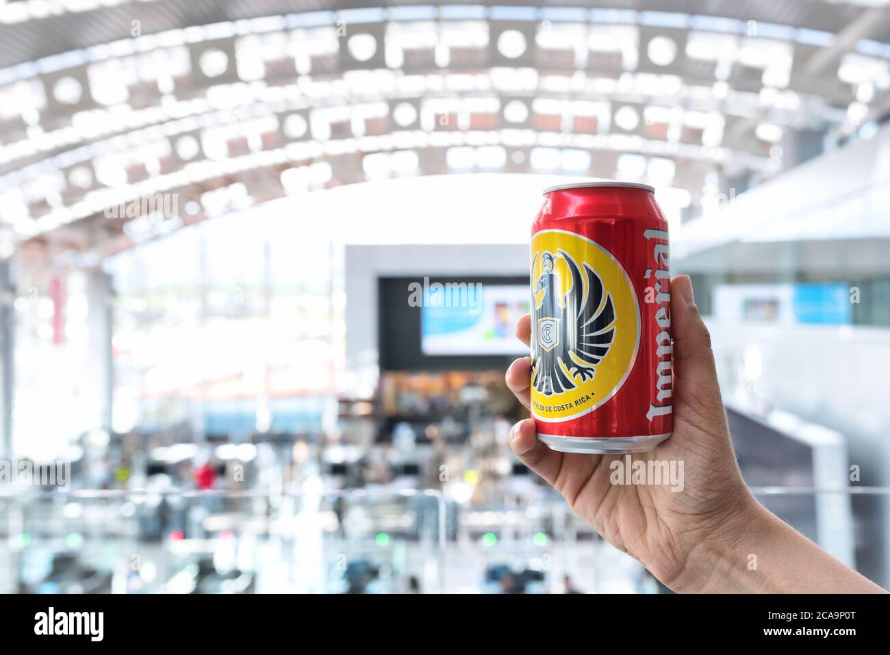 Hand holding an Imperial beer can, the most common beer brand of Costa Rica. Defocused interior view of Juan Santamaría International Airport. Stock Photo